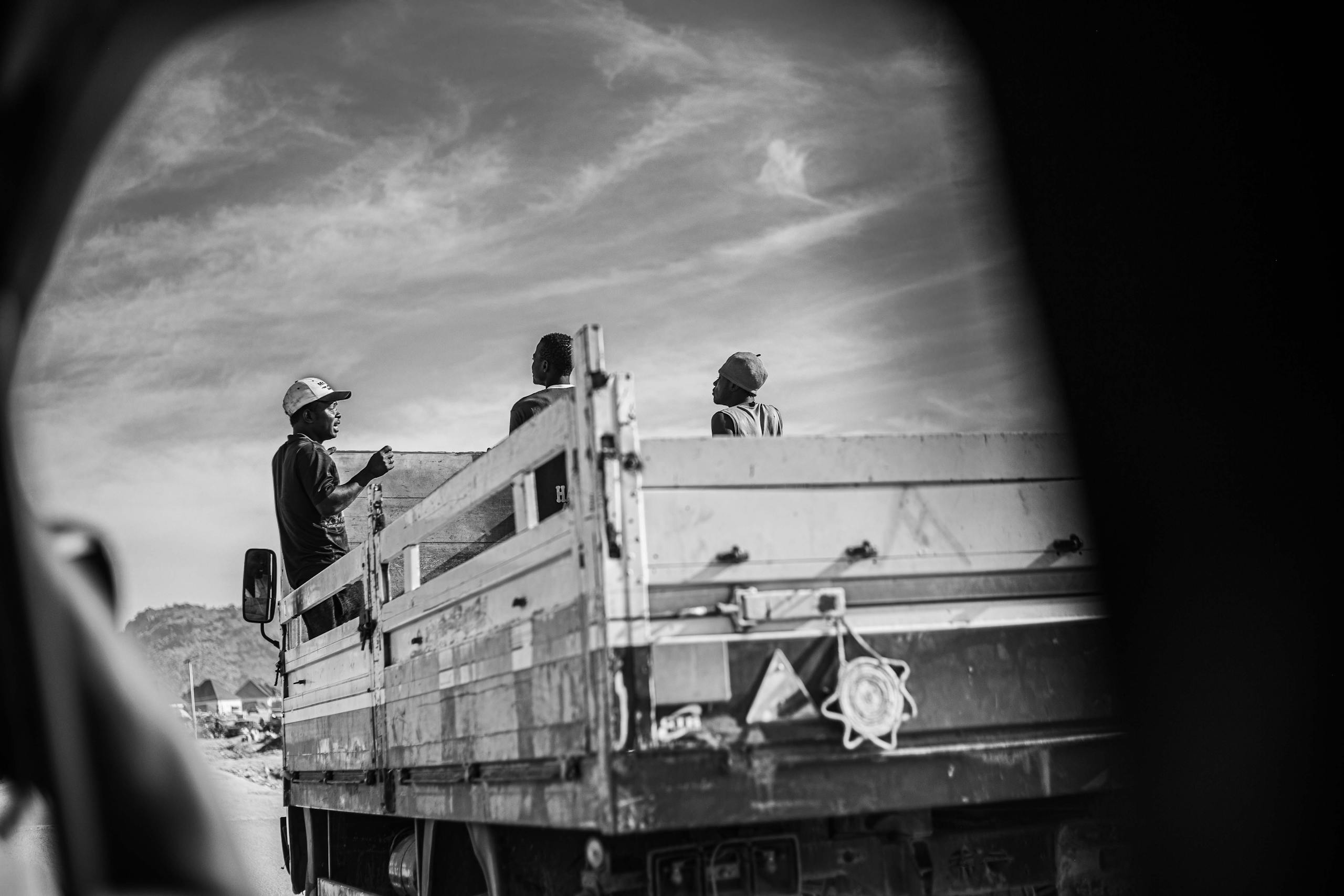 Monochrome image of workers on a truck under blue skies, captured through a vehicle window.